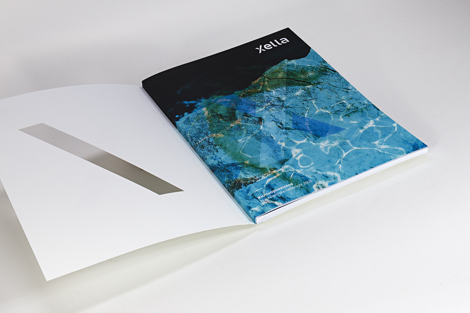Die aufgeschlagene Broschüre der Firma "Xella". Der Umschlag ist weiß, darauf ist eine Linie des großes "X" ausgestanzt. Die erste Seite ist blau-türkis wie Wasser, auf das die Sonne scheint, mit einigen Lichtreflexionen. In kleiner weißer Schrift steht dort: Richtungsweisend. Nachhaltigkeitsbericht 2020 Die aufgeschlagene Broschüre der Firma "Xella". Der Umschlag ist weiß, darauf ist eine Linie des großes "X" ausgestanzt. Die erste Seite ist blau-türkis wie Wasser, auf das die Sonne scheint, mit einigen Lichtreflexionen. In kleiner weißer Schrift steht dort: Richtungsweisend. Nachhaltigkeitsbericht 2020