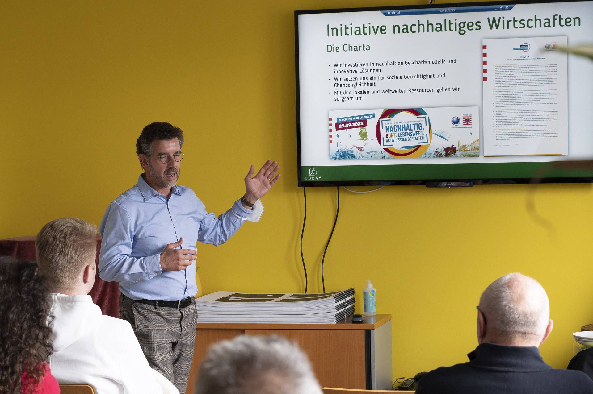 Ralf Lokay steht vor einer Gruppe von Menschen und spricht. Mit einer Hand deutet er auf einen Bildschirm, auf dem ein Slide mit der Überschrift "Initiative nachhaltiges Wirtschaften" zu sehen ist. 
