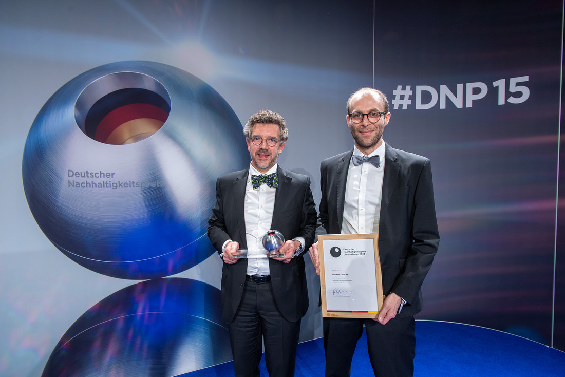 Zwei Männer in schwarzen Anzügen stehen nebeneinander. Einer hält eine Trophäe, die Kugel des Deutschen Nachhaltigkeitspreises in der Hand, der andere eine Urkunde. Im Hintergrund ist ein großes Bild der Deutschen-Nachhaltigkeitspreis-Kugel und der Hashtag #DNP15