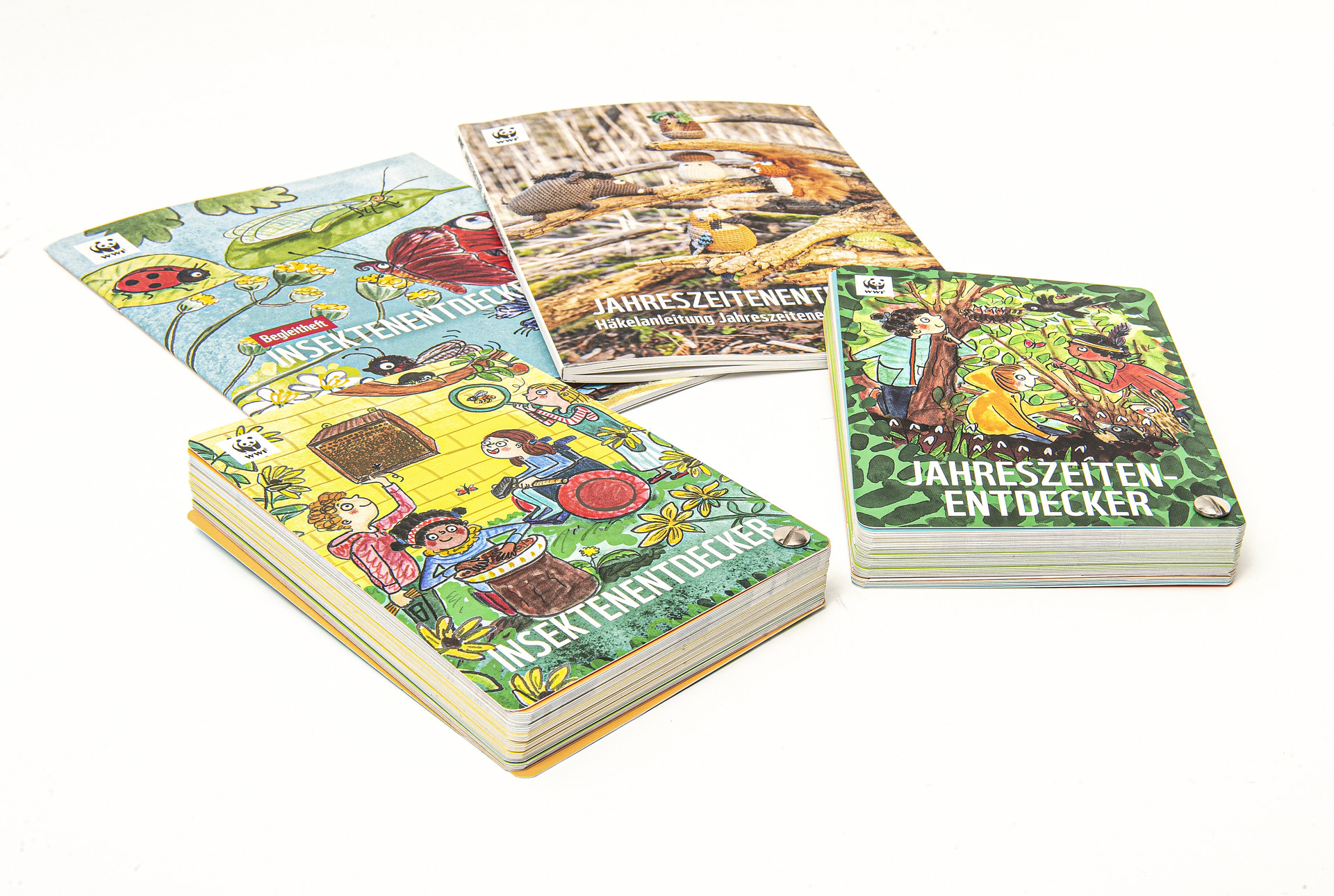 Im Vordergrund liegen zwei Kartensets, im Hintergrund zwei Broschüren. Auf allen Covern sind bunte Illustrationen von Szenen in der Natur. 