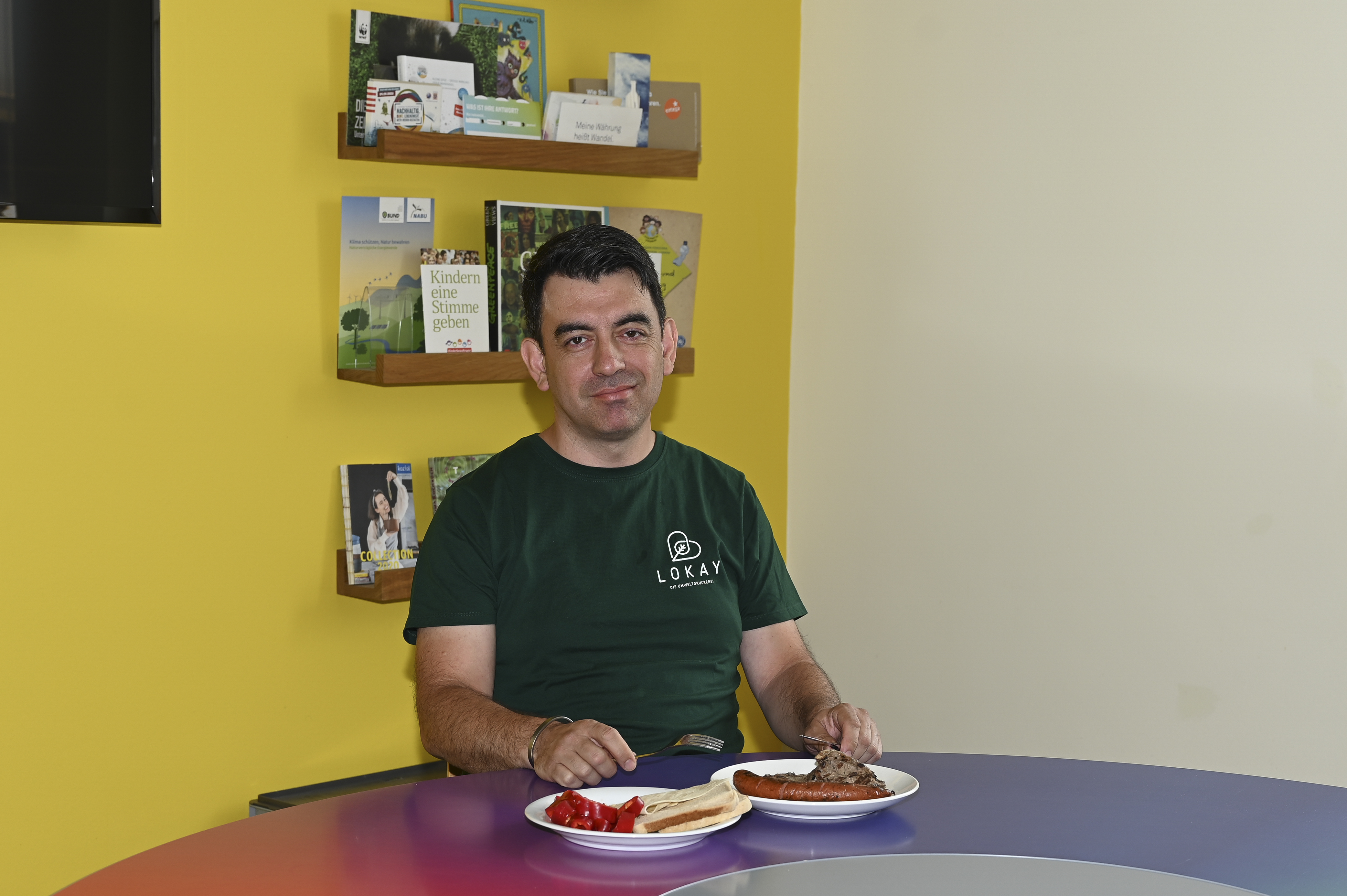Ismail sitzt an einem Tisch und hat zwei Teller vor sich. Er trägt ein grünes T-Shirt mit Lokay-Logo. Hinter ihm ist ein Regal mit Druckmustern zu erkennen. 