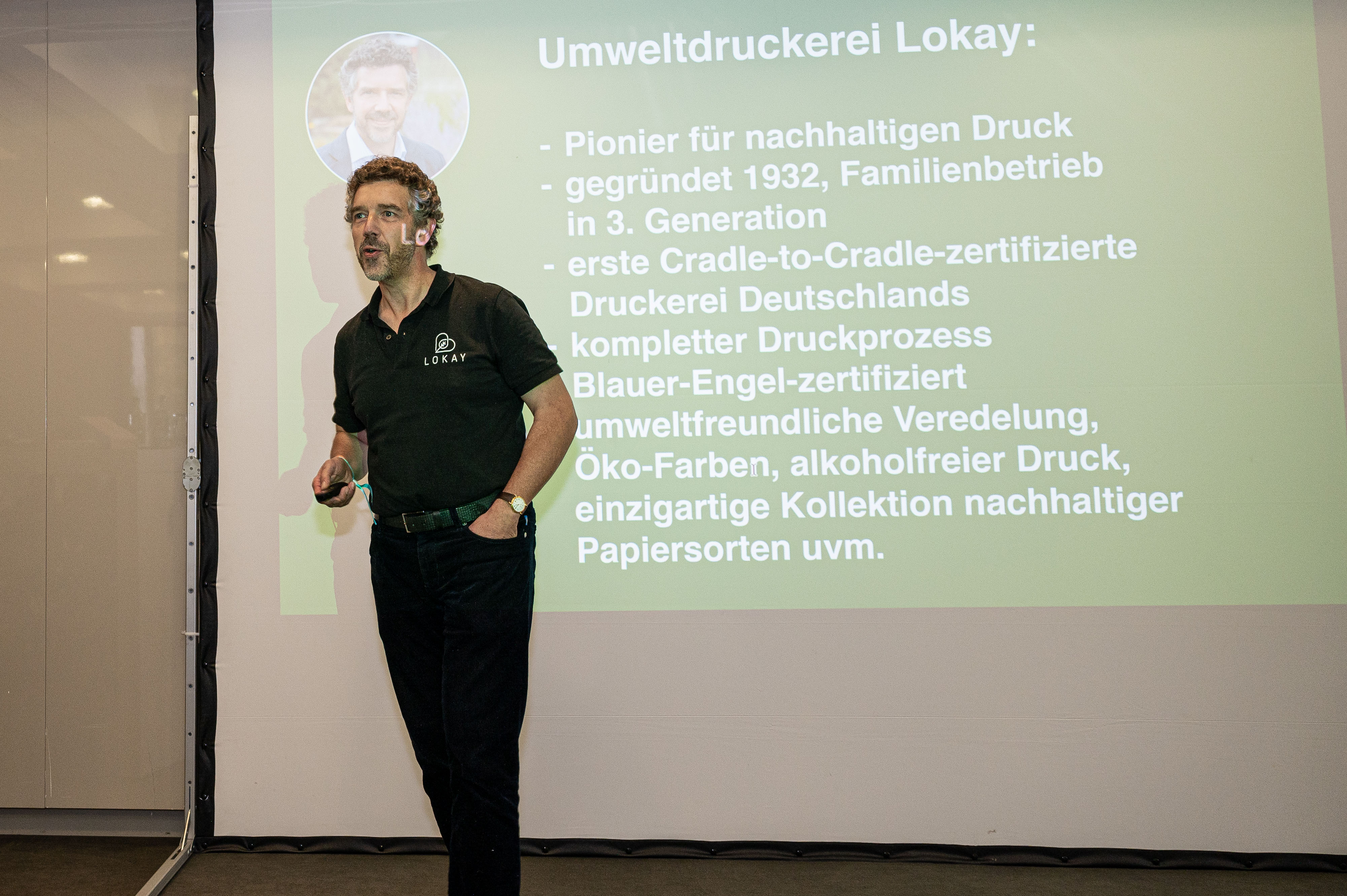 Ralf Lokay steht auf einer Bühne, hinter ihm ist eine große Leinwand, auf der ein Slide mit einer Unternehmensvorstellung von Lokay zu sehen ist. 
