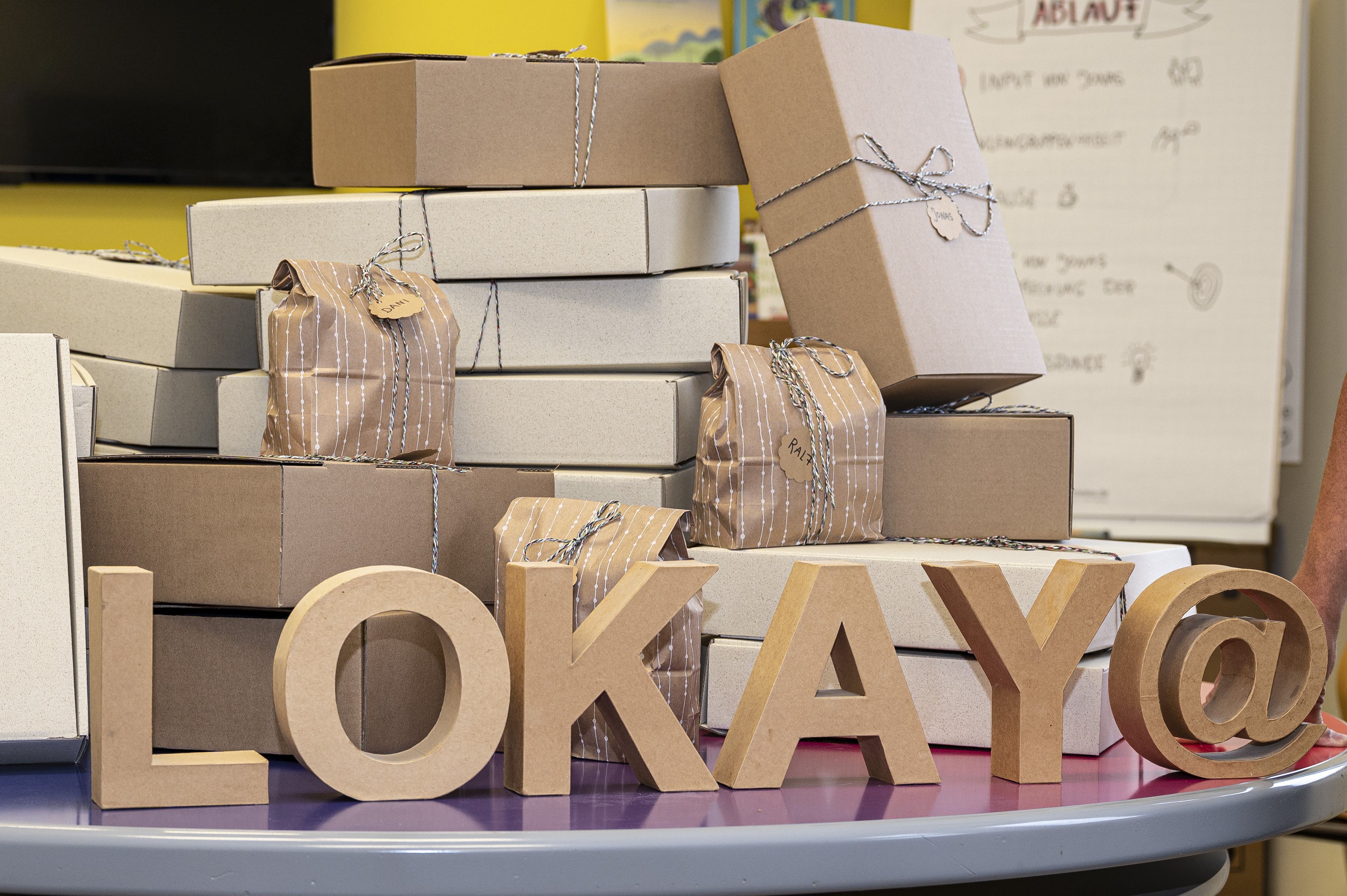 ein Stapel verschieden großer Kartons und Päckchen mit Schleifen. Im Vordergrund stehen Papp-Buchstaben, die das Wort "LOKAY" bilden