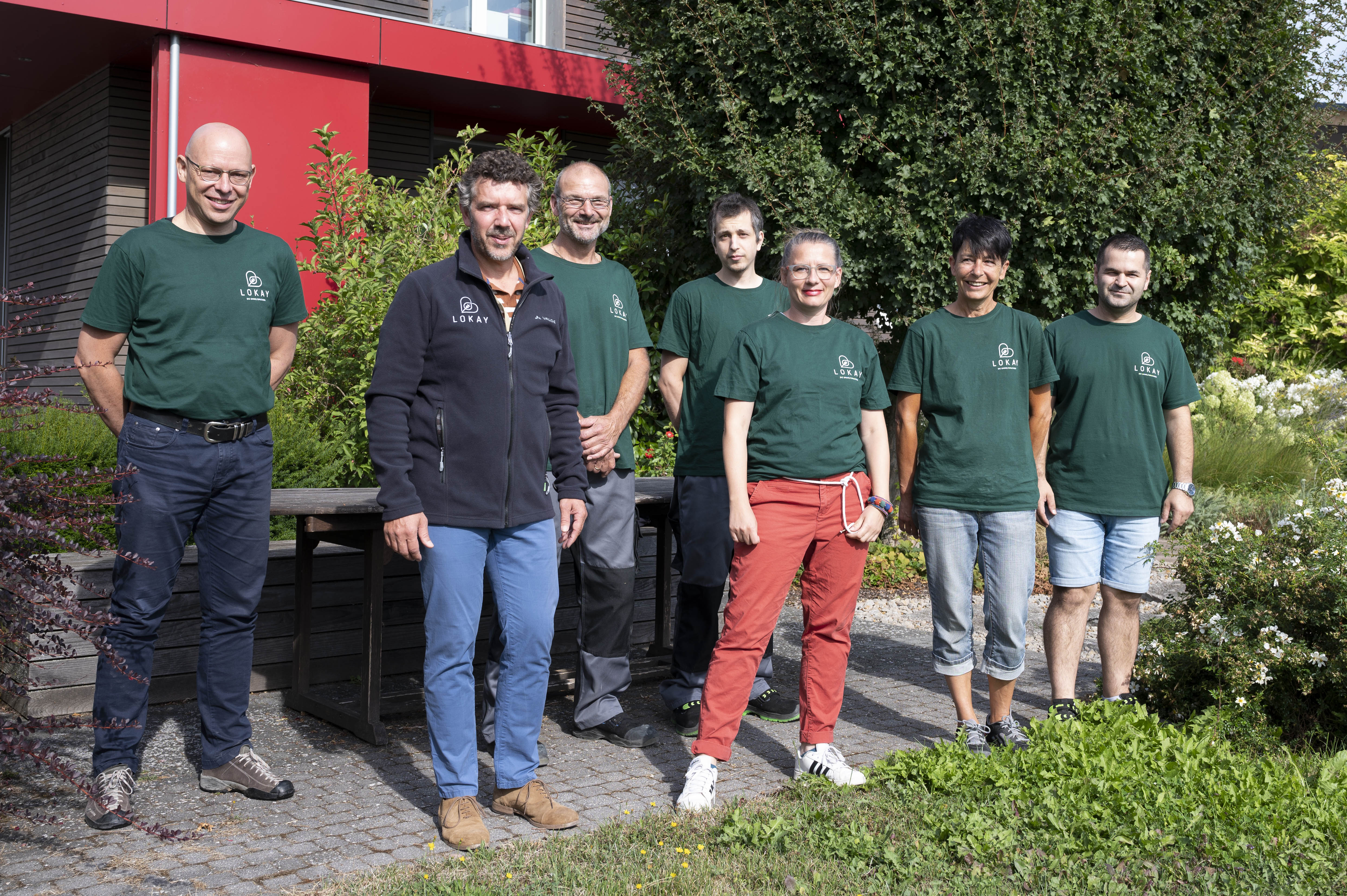 Sechs Personen stehen mit den neuen dunkelgrünen Tshirts im Firmengarten. Die Tshirts tragen ein kleines Lokay-Logo in weiß auf der linken Brust. Ralf Lokay steht in mitten der Leute. Er trägt die passende, schwarze Fleece-Jacke, ebenfalls mit Lokay-Logo auf der Brust. 