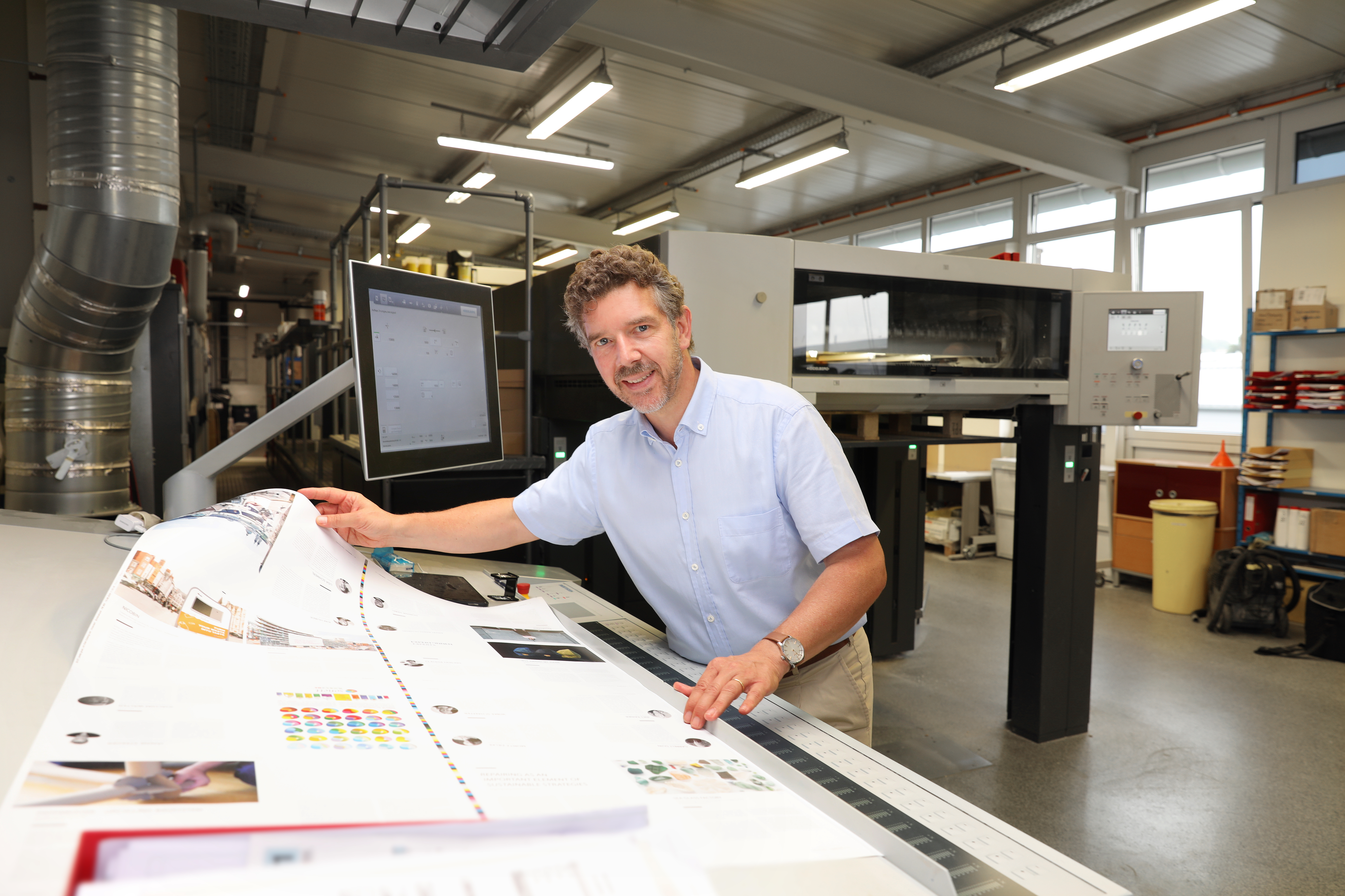 Ralf Lokay steht in der Produktionshalle an einem Tisch. Er hält einen Bogen Papier in der Hand und lächelt in die Kamera.